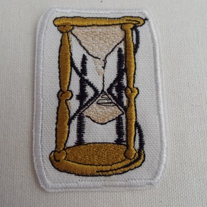 May include: A white embroidered patch with a gold hourglass design. The hourglass is filled with sand and has a black outline.