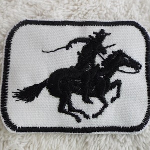 Puede incluir: Parche bordado negro de un vaquero montando un caballo, con fondo blanco y borde negro.