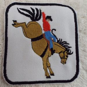 Peut inclure: Patch brodé représentant un cowboy chevauchant un cheval brun qui se cabre. Le cowboy porte une chemise rouge, un jean bleu et un chapeau marron. Le cheval se cabre, ses pattes avant en l'air. Le patch est bordé de noir.