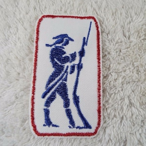 May include: A rectangular embroidered patch featuring a white background with a red stitched border. The center displays a dark blue silhouette of a soldier in a tricorn hat holding a long rifle, standing in a profile position facing right.