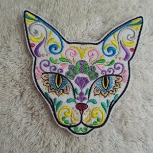 May include: Embroidered iron-on patch of a white cat's face with colorful Day of the Dead inspired designs. The cat's eyes are blue, green, and yellow.