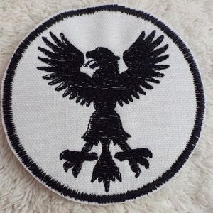 May include: A white circular patch with a black embroidered eagle design. The eagle has its wings spread and is facing to the right.