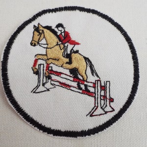 May include: A white embroidered patch with a black border depicting a brown horse jumping over a red and white fence with a rider in a red jacket.