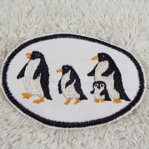 May include: Embroidered patch featuring a penguin family. The patch has a white background with a black oval border. The penguins are black and white with yellow beaks and feet. The family consists of three adult penguins and one baby penguin.