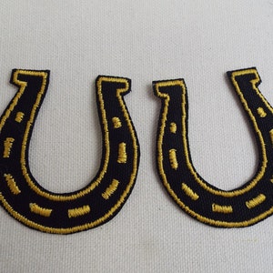 May include: Two black fabric horseshoe patches with gold stitching. The horseshoe patches are about 2 inches tall.