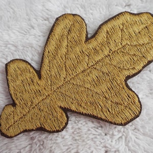 May include: A gold embroidered oak leaf patch. The leaf is detailed with stitching and has a brown felt backing.