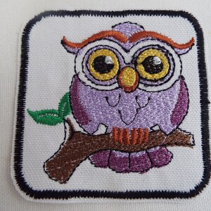 May include: Purple embroidered owl perched on a brown branch with green leaves. The owl has large yellow eyes and orange eyebrows. The owl is surrounded by a black border.