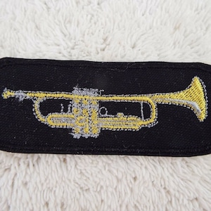 Könnte beinhalten: Ein schwarzer gestickter Aufnäher mit einem goldenen Trompetenmotiv.