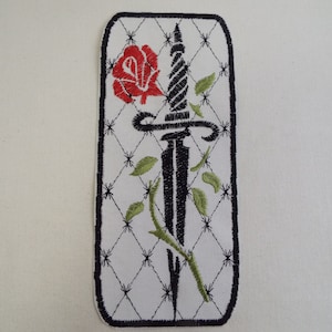 Puede incluir: Parche bordado en negro y rojo con una daga y una rosa, rodeado de un fondo acolchado blanco.