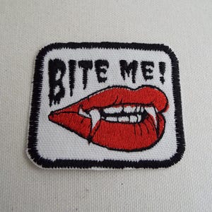 May include: White embroidered patch with black stitching. The patch features a red, open-mouthed, vampire-like lip with fangs and the text "Bite Me!" in black.