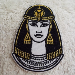 May include: Embroidered patch featuring a stylized depiction of an Egyptian queen. The patch has a black background with gold and white details, including a cobra headdress, earrings, and a collar. The patch is approximately 3 inches tall.