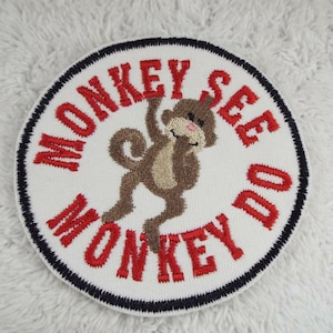 May include: A white, round embroidered patch with a red border. The patch features a brown monkey with the text "Monkey See Monkey Do" in red.