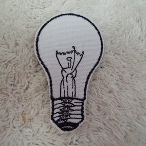 Może przedstawiać: Biała, haftowana naszywka w kształcie żarówki. Żarówka ma czarny kontur i szczegółowy czarny wzór żarnika wewnątrz. Naszywka znajduje się na białej, teksturowanej powierzchni.