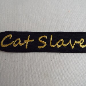 May include: Black fabric patch with gold embroidered text that reads "Cat Slave".