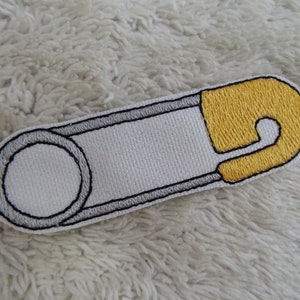 Puede incluir: Parche de planchado blanco bordado de un imperdible con la parte superior amarilla. El imperdible está contorneado en negro y gris.