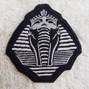 May include: A black fabric patch with a white embroidered image of the pharaoh's head, known as the Sphinx, with a crown on top.