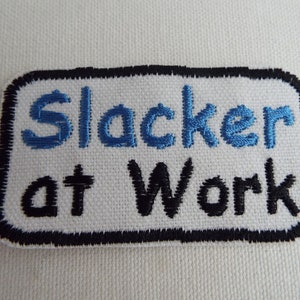 May include: White embroidered patch with black stitching around the edges. The patch says "Slacker at Work" in blue and black thread.