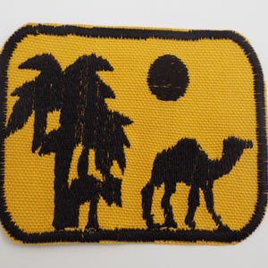May include: A rectangular embroidered patch with rounded corners featuring a yellow background and a black border. The design depicts a black silhouette of a palm tree on the left, a solid black circle representing the sun, and a camel.