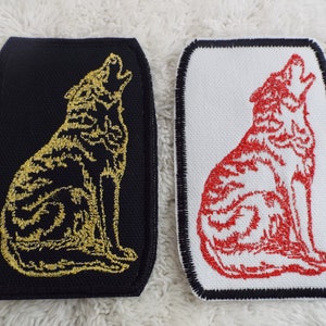 May include: Two rectangular patches featuring a howling wolf design. One patch is black with a gold embroidered wolf, the other is white with a red embroidered wolf. Both have a dark border.