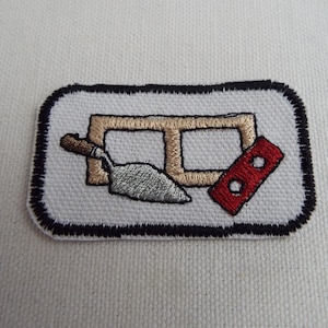 Puede incluir: Un parche bordado blanco con un borde negro. El parche presenta un ladrillo marrón con dos agujeros, una paleta plateada y un ladrillo rojo con dos agujeros.