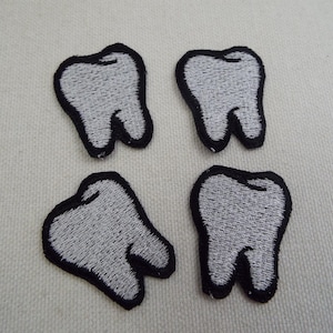 May include: Four embroidered patches in the shape of teeth. Each tooth is white with black outlining and details. The patches are arranged on a light-colored fabric background.