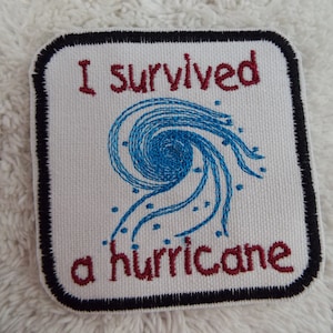 May include: White embroidered patch with a black border. The patch features a blue hurricane symbol and the text "I survived a hurricane" in red.