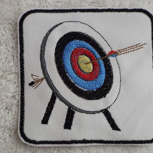 May include: Embroidered patch featuring a white archery target with black, blue, red, and yellow rings. An arrow with brown fletching is centered in the target. The target is on a black stand with three legs.