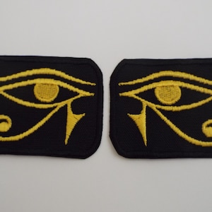 Può includere: Due toppe nere ricamate con il simbolo dell'Occhio di Horus in giallo. Le toppe sono rettangolari con angoli arrotondati. Il disegno dell'occhio comprende un occhio stilizzato, un sopracciglio e segni.