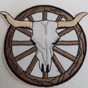 May include: Embroidered patch featuring a white bull skull with brown horns, centered within a brown wagon wheel with spokes.