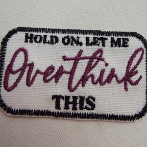 May include: White rectangular embroidered patch with black stitching around the edges. The text "Hold on, let me overthink this" is embroidered in black and purple.