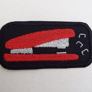 Op de afbeelding: Rode en witte geborduurde strijk-op patch van een nietmachine met een zwarte achtergrond.