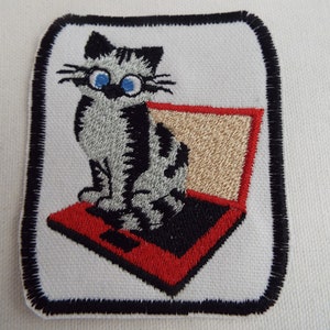 Peut inclure: Patch thermocollant brodé représentant un chat de dessin animé assis sur un ordinateur portable rouge. Le chat a une fourrure grise et noire, des yeux bleus et des moustaches blanches.