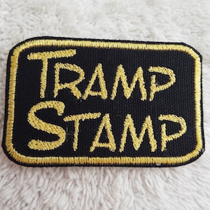 Könnte beinhalten: Ein schwarzer gestickter Aufnäher mit goldenen Nähten, auf dem "TRAMP STAMP" steht.