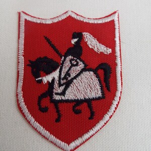 Puede incluir: Un parche bordado rojo en forma de escudo con un borde blanco. El parche presenta un caballero blanco montando un caballo negro con una silla blanca y una pluma blanca en el casco del caballero.