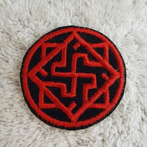 May include: A round black patch with a red embroidered design. The design is a stylised cross with a geometric pattern around it.