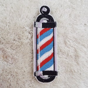 May include: A red, white, and blue barber pole with a black base and a silver top. The barber pole is embroidered on a white fabric.