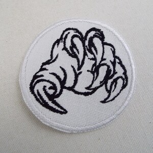 May include: White embroidered patch featuring a black claw design. The claw is shown in a grasping pose with sharp, pointed nails.
