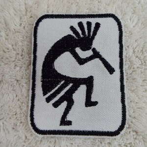 May include: A rectangular patch featuring a black Kokopelli figure playing a flute on a white background. The patch has a black border and is likely made of fabric.