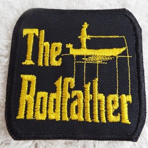 May include: A black fabric patch with yellow embroidered text that reads "The Godfather" and a silhouette of a boat with a person standing on it.