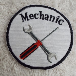 May include: White embroidered patch with a black border. The patch features a wrench and screwdriver crossed over each other. The word "Mechanic" is embroidered in black above the tools.