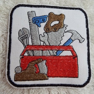Puede incluir: Un parche blanco bordado con una caja de herramientas roja llena de herramientas. Las herramientas incluyen un martillo, una sierra, una llave inglesa, una regla y un cepillo. El parche tiene un borde negro.