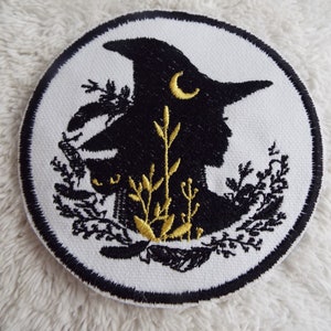 May include: A white embroidered patch with a black silhouette of a witch wearing a hat with a crescent moon on it. The witch is surrounded by gold and black floral designs.