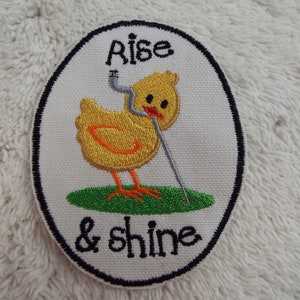 Könnte beinhalten: Gestickter Bügel-Patch mit einem gelben Küken, das einen Golfschläger hält. Der Patch ist oval geformt mit weißem Hintergrund und schwarzem Rand. Der Text "Rise & Shine" ist in Schwarz gestickt.
