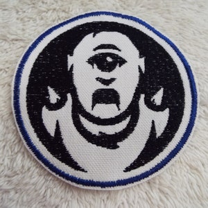 Peut inclure: Un écusson brodé blanc avec un contour noir et une bordure bleue. L'écusson représente une image stylisée d'un visage avec un seul grand œil et une moustache.