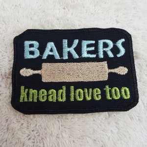 May include: Black embroidered patch with the text "BAKERS knead love too" in green and blue lettering. A beige rolling pin is embroidered in the center of the patch.