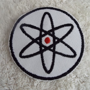 May include: A white embroidered patch with a black atom symbol and a red dot in the centre.