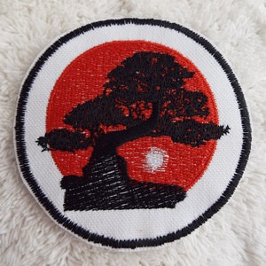 May include: A white embroidered patch with a black silhouette of a bonsai tree in front of a red sun. The patch has a black border.