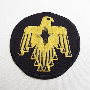 Può includere: Una toppa rotonda nera con un designo di uccello stilizzato ricamato in giallo. L'uccello ha un occhio nero e una forma di diamante nero al centro del petto.