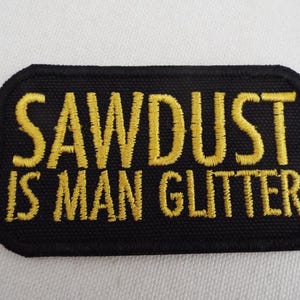 Puede incluir: Parche rectangular negro con esquinas redondeadas. El parche presenta el texto bordado "SAWDUST IS MAN GLITTER" en amarillo. El parche es probablemente para ropa o accesorios.