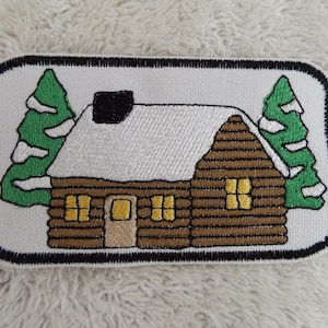 Può includere: Una toppa ricamata che rappresenta un cottage marrone con un tetto innevato e due alberi sempreverdi. Il cottage ha un camino nero e finestre con luci gialle.